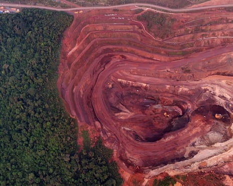 Aerial view of a large open-pit iron ore mine with terraced red earth excavations beside dense rainforest