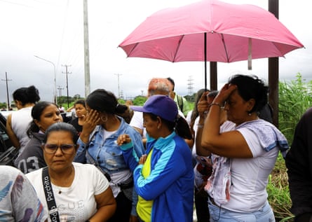 Women gather on a roadside. One woman is clutching her head while carrying a pink umbrella