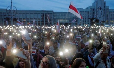 A large crowd holding up phones with the torch function illuminated, with a white and red Belarusian flag in the background.
