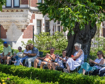 A row of people sitting on chairs under the shade of a tree.