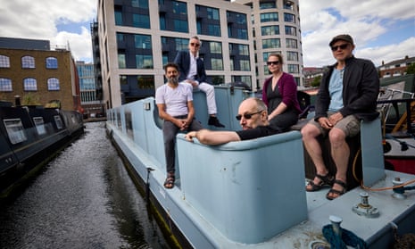 four men and a woman on the deck of a pale blue steel narrowboat; one man is sitting on the cabin roof and another on the edge of the guard rail while the others are on benches along the side. Other boats can be seen either side plus a large modern office building in the background across the canal basin.