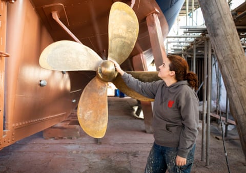 A woman touches a metre-wide brass propeller in a boatyard