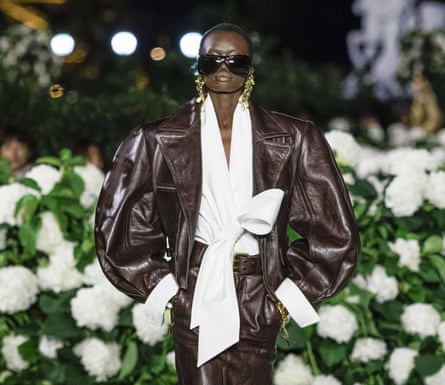 Model wearing white blouse with large pussy bow, beneath a brown leather jacket with voluminous sleeves
