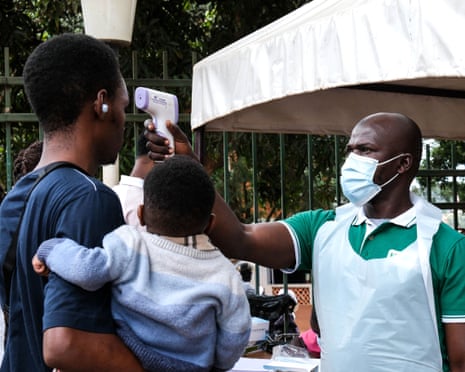 A person in a surgical mask and plastic apron holds a thermometer to the head of a man carrying a child.
