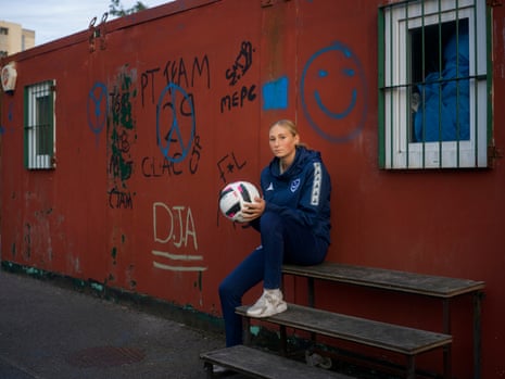A teenage girl in a tracksuit sits on a bench holding a football