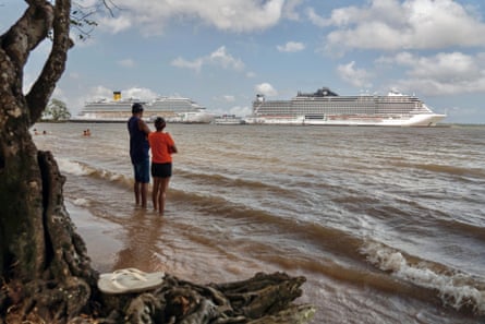 People stand in the shallows of a river looking out at cruise ships