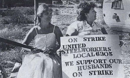 Two strikers’ wives sit on the picket line holding a banner showing support for their husbands in a black and white picture from the strike.