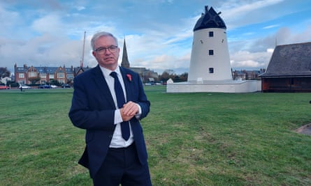 Mark Menzies: he is wearing a dark suit, white shirt and tie and is seen standing on grass in front of a white windmill