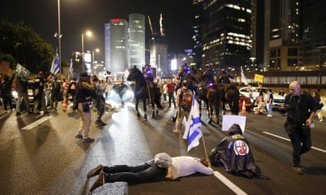 People protest on a road in Tel Aviv at night, with tower block behind. One person lies on the ground holding an Israeli flag