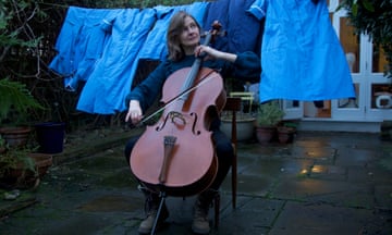 A woman plays a cello in a garden in front of a washing line hung with blue nurses' uniforms