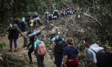 A long, close line of people carrying backpacks trudges along a muddy jungle trail.