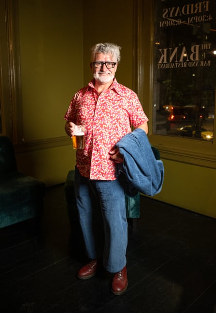 A man in blue corduroy pants and a red and white button short-sleeve shirt, holding a glass of beer, with a blue corduroy jacket draped over his arm. He looks happy with himself.