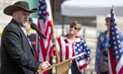 A man wearing a black suit and a brown cowboy hat speaks in front of a podium with American flags in the background.