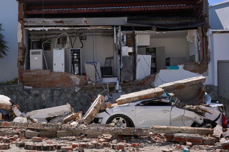 A crushed silver car is covered in fallen concrete outside a destroyed building.