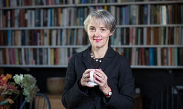 Lucy Kellaway, with short hair and wearing a watch and a necklace with round beads under a suit jacket, smiles as she holds a mug in front of a bookcase