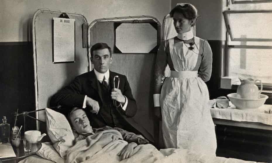 Dr Basil Hood sits by the bedside of a patient as a nurse watches on.