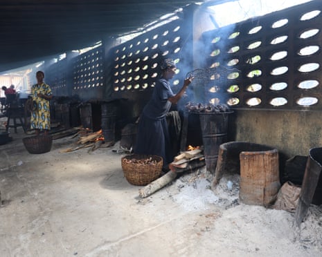 ‘The water is no longer our friend’: how dredging is pushing Lagos Lagoon towards ecosystem collapse – photo essay A woman smokes fish over a stove made from a oil drum in a shed with other fish smokers.