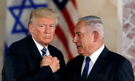 two men in suits grasp each other's hand in front of US and Israeli flags