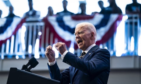 An older white man in a dark suit and tie speaks in an emphatic manner, with both fists balled an raised, from behind a lectern. Behind and above him is a railing adorned with red-white-and-blue bunting, which listeners lean upon.