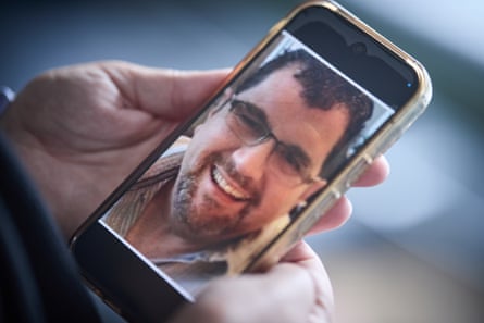 Adrian Howe pictured on the screen of his daughter’s phone, which she is holding in her hands. He is in his 50s with short dark hair, a short beard and glasses, and smiling widely in this image.
