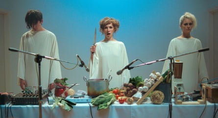 film still of three people in white standing behind a table laid with lots of food, with microphones in front