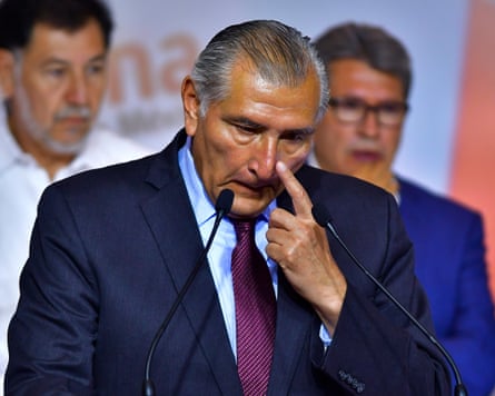 An older man in a suit and dark purple tie touches his nose as he stands in front of two microphones.