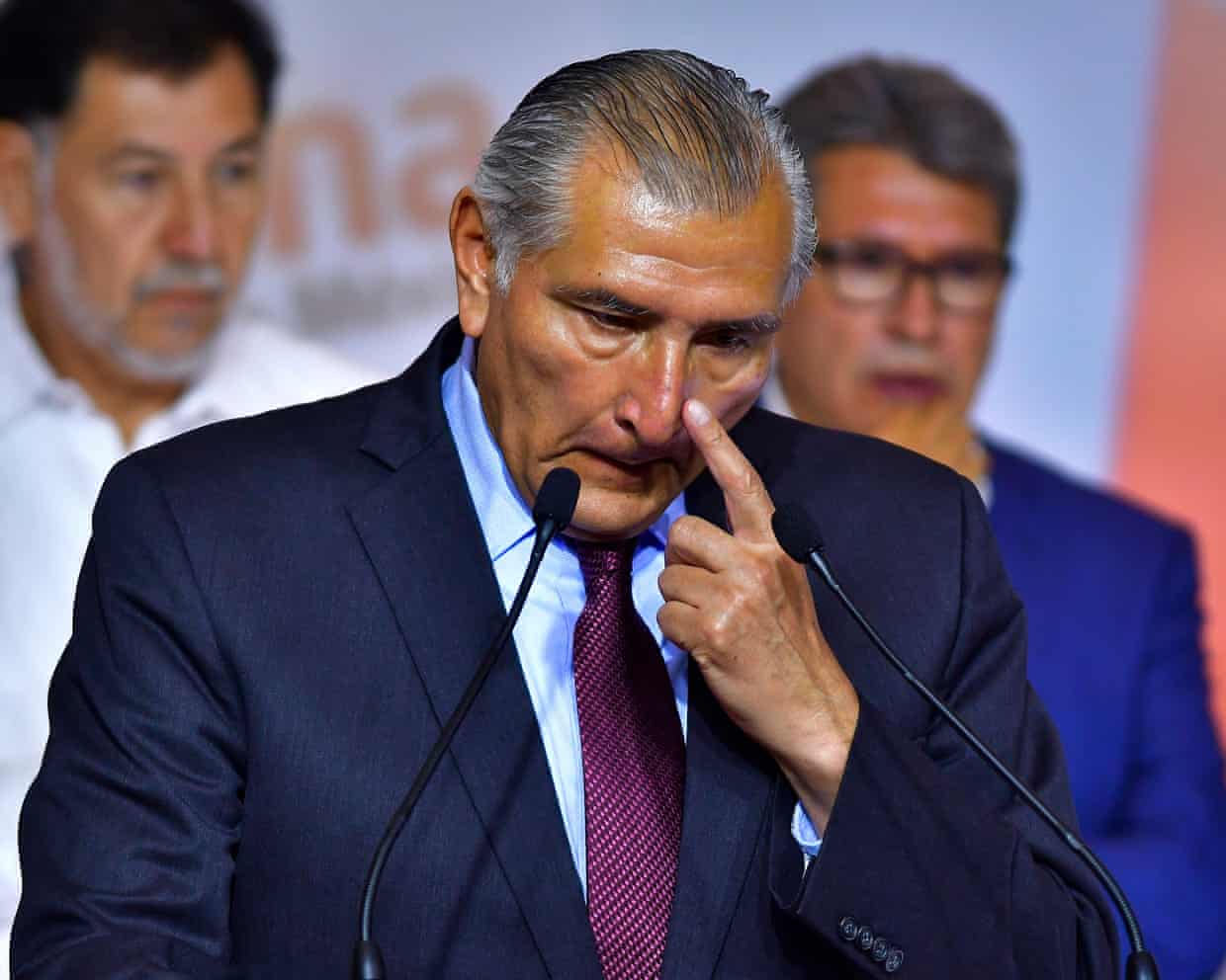 An older man in a suit and dark purple tie touches his nose as he stands in front of two microphones.