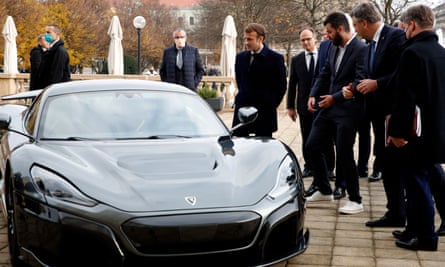 Macron, in an overcoat, looks at a swoopy black supercar parked outside on a terrace, with a group of bystanders including Mate Rimac