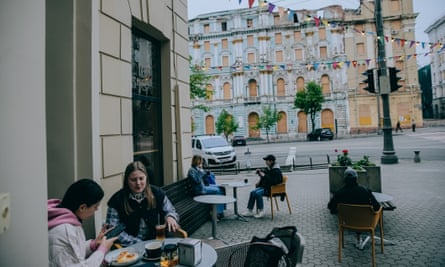 People sitting outside a cafe with a boarded-up building across the road.