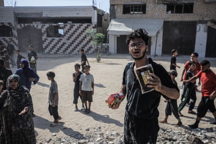 A man holds packets of what appears to be food. Other people can be seen on the dusty street behind him