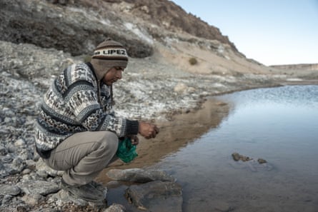 A man crouches by a lake in front of a steep rocky slope