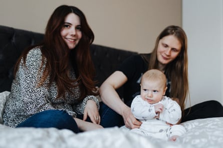 A woman with long dark hair and a man with long fair hair sit on a bed with a baby in a sleepsuit