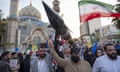 A model of a hypersonic missile is carried past a mosque, seen in the background, by a group of people who have their arms raised; some look jubilant and there is a large green, white and red Iranian flag billowing