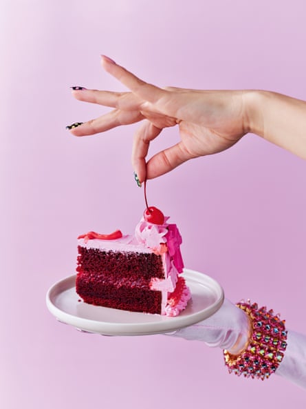 A slice of red velvet cake with pink buttercream and raspberry compote filling with a cherry on top, on a pink background