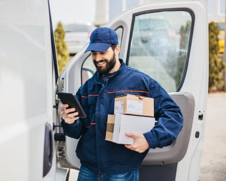 A delivery man in a navy jacket and baseball cap with parcels in his hand stands next to his van.