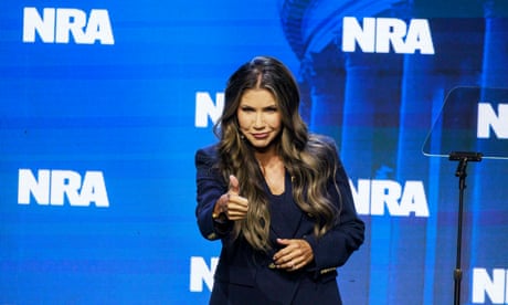 a woman smiles and gives a thumbs up while standing on stage in front of multiple logos for the national rifle association