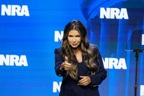 a woman smiles and gives a thumbs up while standing on stage in front of multiple logos for the national rifle association