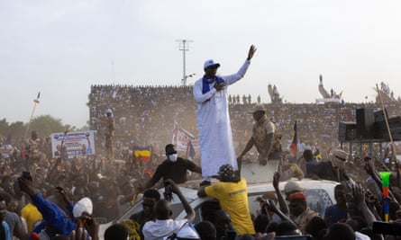 A man stands on top of a car at an election campaign rally and, surrounded by a busy people who appear to be waving and cheering, he waves to his supporters
