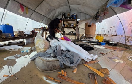 A man stands in ankle-deep water inside a flooded tarpaulin shelter; a sodden mattress piled with wet possessions is in the foreground and bits of wood, rugs and various containers float in the muddy water.