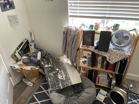 a messy room with a pile of posters and other papers in the corner, next to a dog bed and a bookcase with a cloth draped over it and a fan, speaker and photo sitting on it