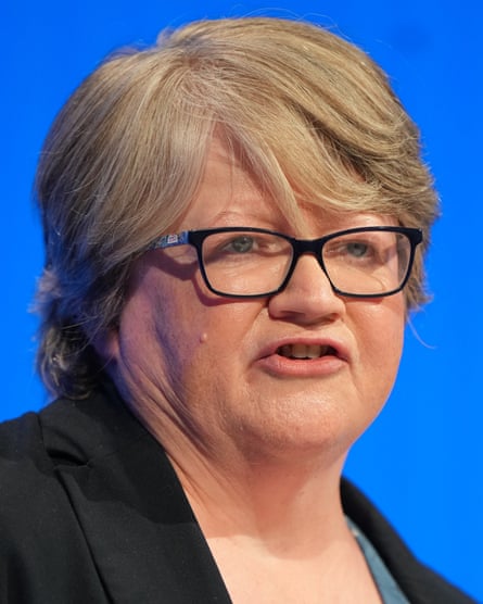 Head shot of Thérèse Coffey against a blue background, wearing a black jacket and glasses, with serious expression, in the middle of giving a speech.
