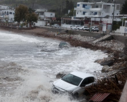 Two cars lying on the shore of a rocky beach with waves crashing over them and houses in the background