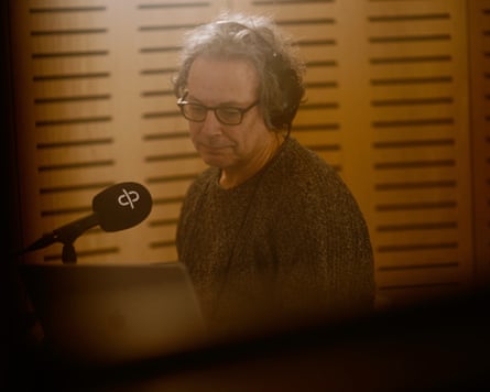 A man with glasses and headphones sits in a booth in front of a microphone.