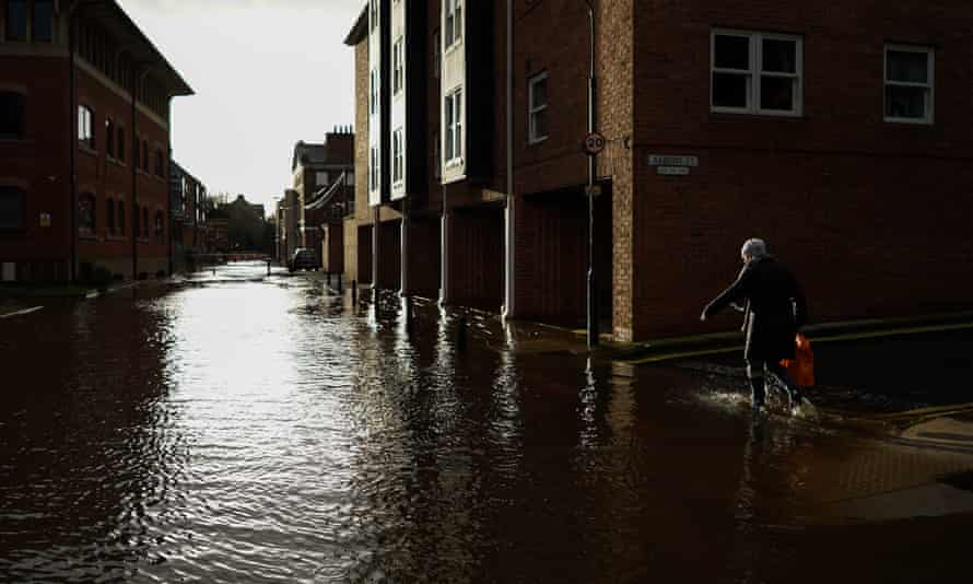 A heavily flooded street illuminated by weak sunlight, with a woman to the right of the picture, wading through water, emerging into the sunlght