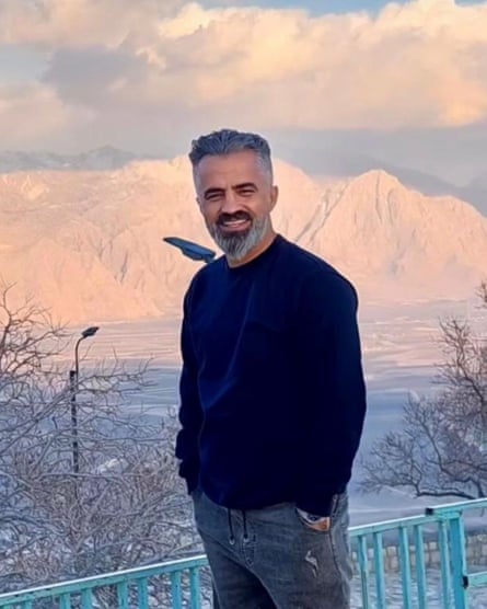 A smiling middle-aged man with a short beard and slicked hair posing outdoors for a picture with mountains in the background