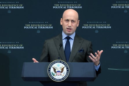 bald man wearing grey suit and navy tie gestures while speaking behind podium