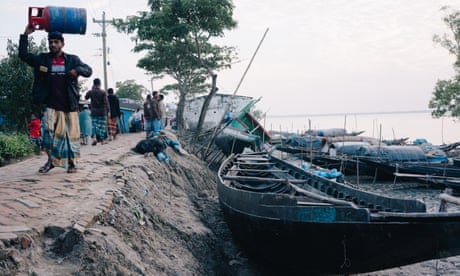 People walk along a raised mud embankment with small boats tied up alongside