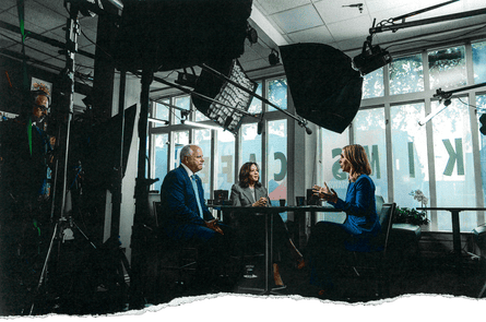 three people speak around a table surrounded by camera equipment
