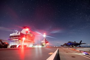 A night shot of an illuminated aircraft carrier flightdeck with fighter jets parked on one side of it