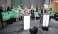 Carla Denyer and Adrian Ramsay standing behind lit-up lecterns with a crowd of supporters behind them with ‘Vote Green’ placards
